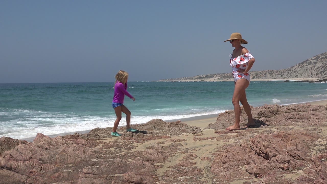 At 0:00 in Cabo San Lucas, a young girl in a purple top approaches a woman in a sun hat on reddish coastal rocks, with surf, sand, and hills behind them.