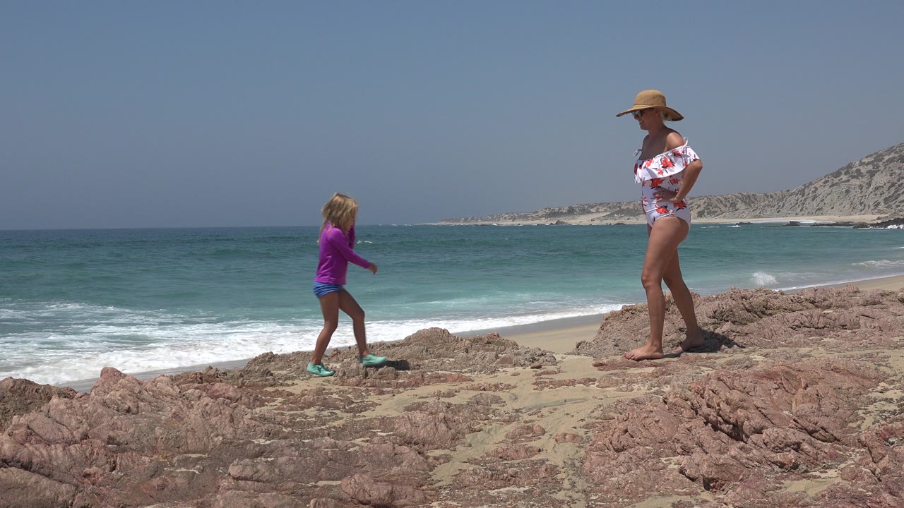 At 0:01 in Cabo San Lucas, the girl steps farther across the uneven rocks toward the woman, while a band of turquoise water and white surf fills the background.