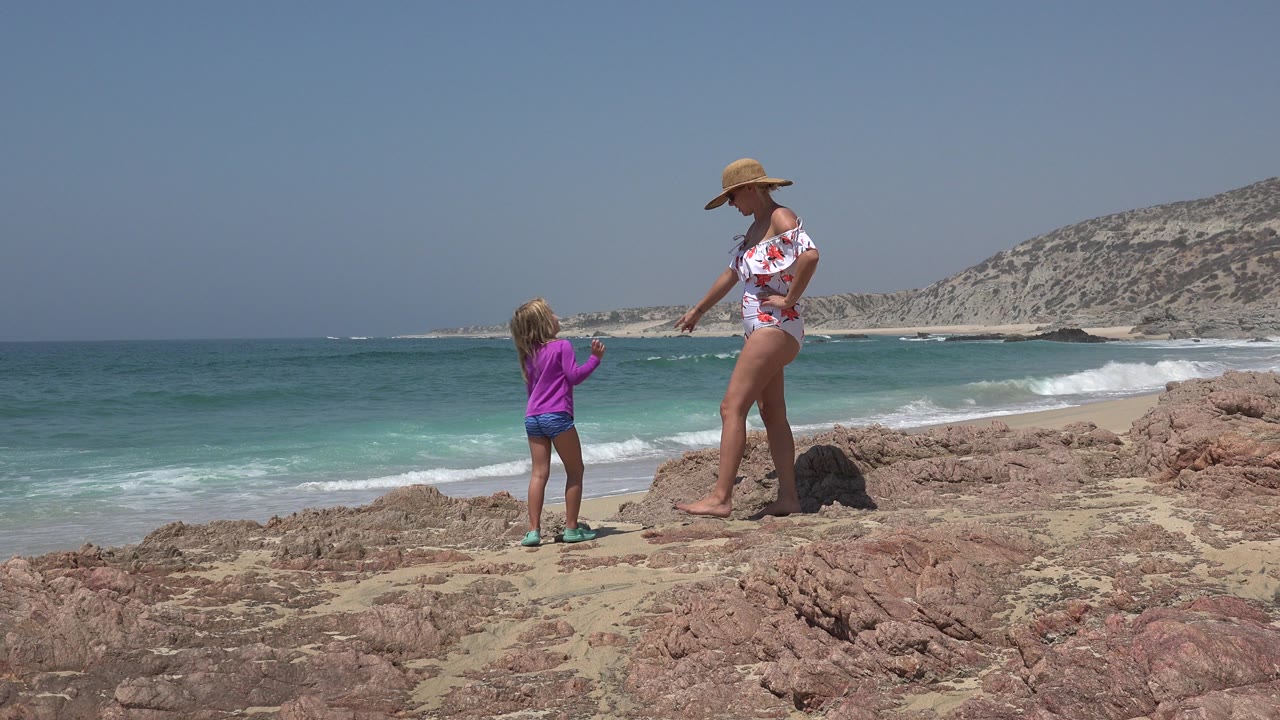 Mother and young daughter standing on beach rocks near ocean surf.