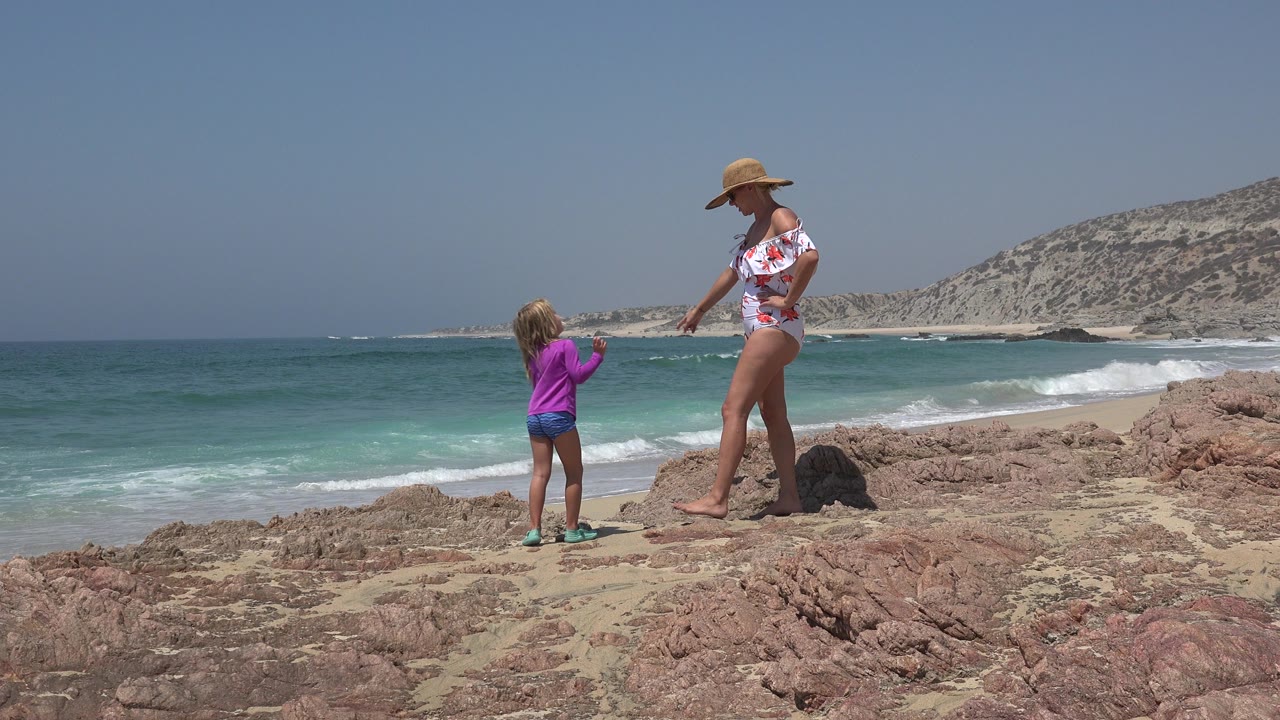 The mother and daughter remain near the same rocky foreground in Cabo San Lucas, Mexico, with the woman turned toward the child and white surf rolling onto the beach behind them.