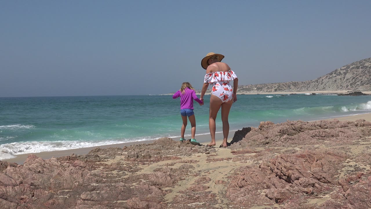 Woman and child stand apart on beach rocks with ocean behind them.