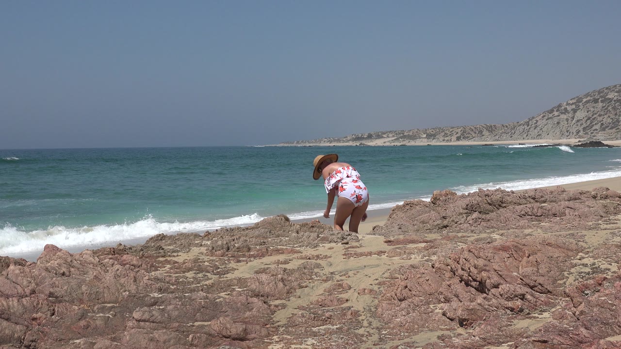 Woman in a straw hat bending on rocks beside the ocean.