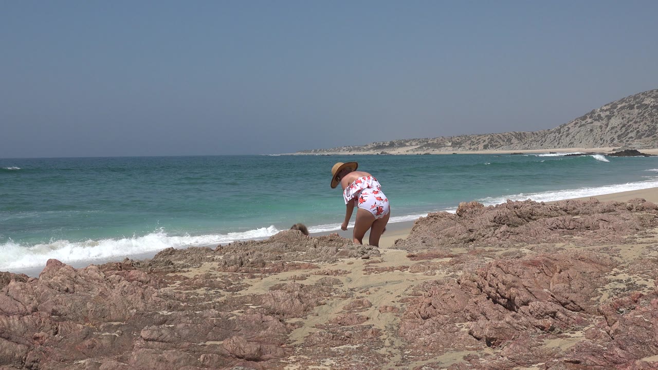 The woman remains crouched on the Cabo San Lucas rocks as white surf rolls in across the turquoise water behind her.
