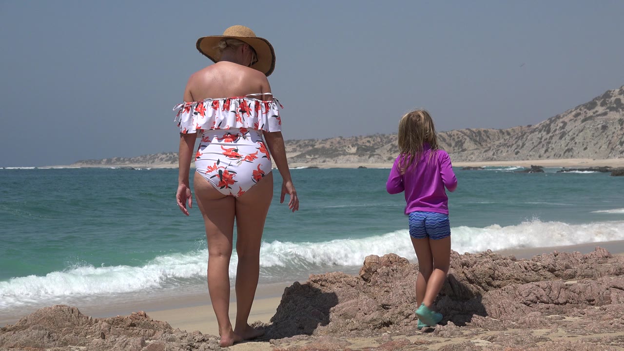 Mother and daughter stand on rocks facing ocean waves.