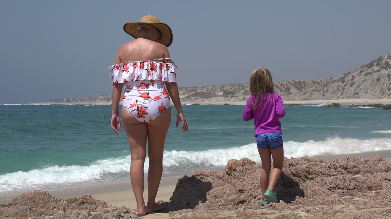 The mother and daughter remain turned away from camera in Cabo San Lucas, with a white wave breaking near shore behind the rocky foreground.