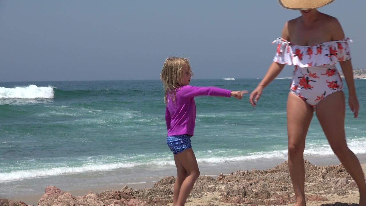 Child points toward an adult woman beside waves on a rocky beach.