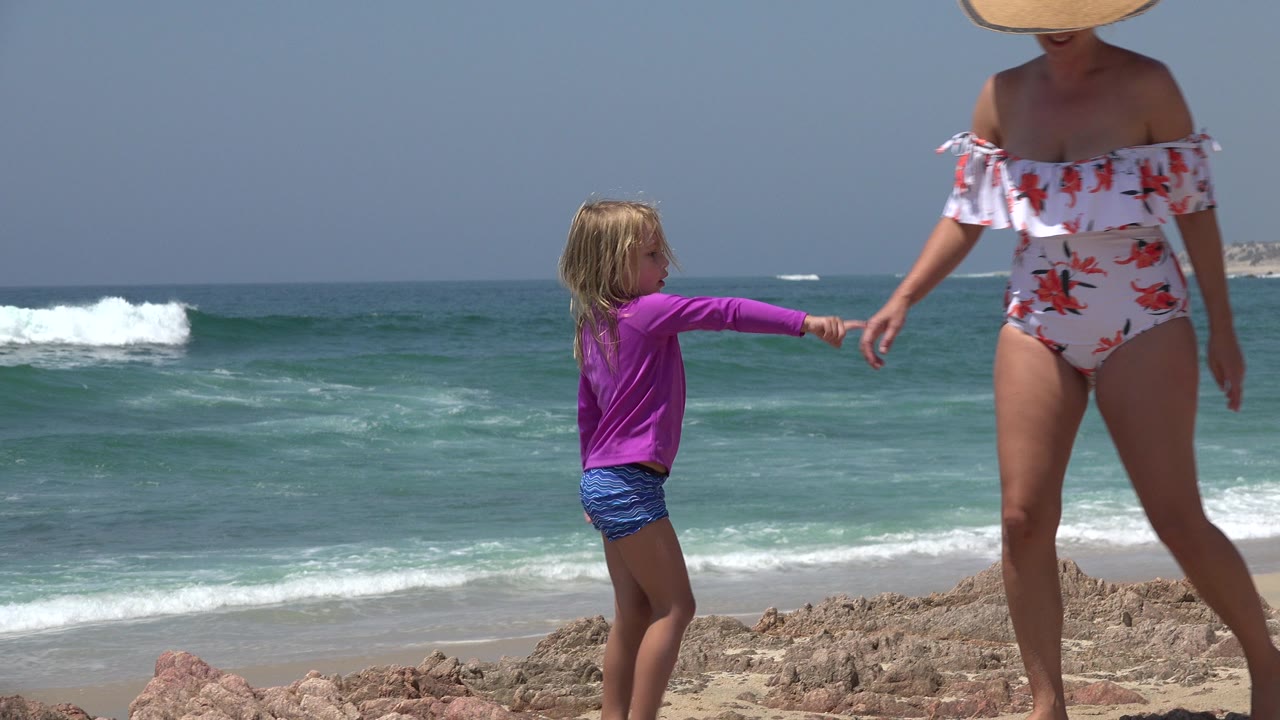 In Cabo San Lucas, Mexico, the child reaches an arm toward the adult woman while standing on rocks, with white surf rolling across the background.