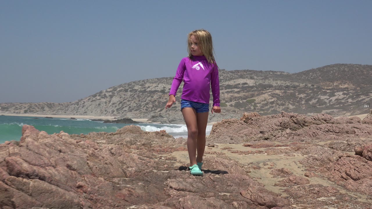 Young girl walking on reddish beach rocks with ocean and hills behind her.