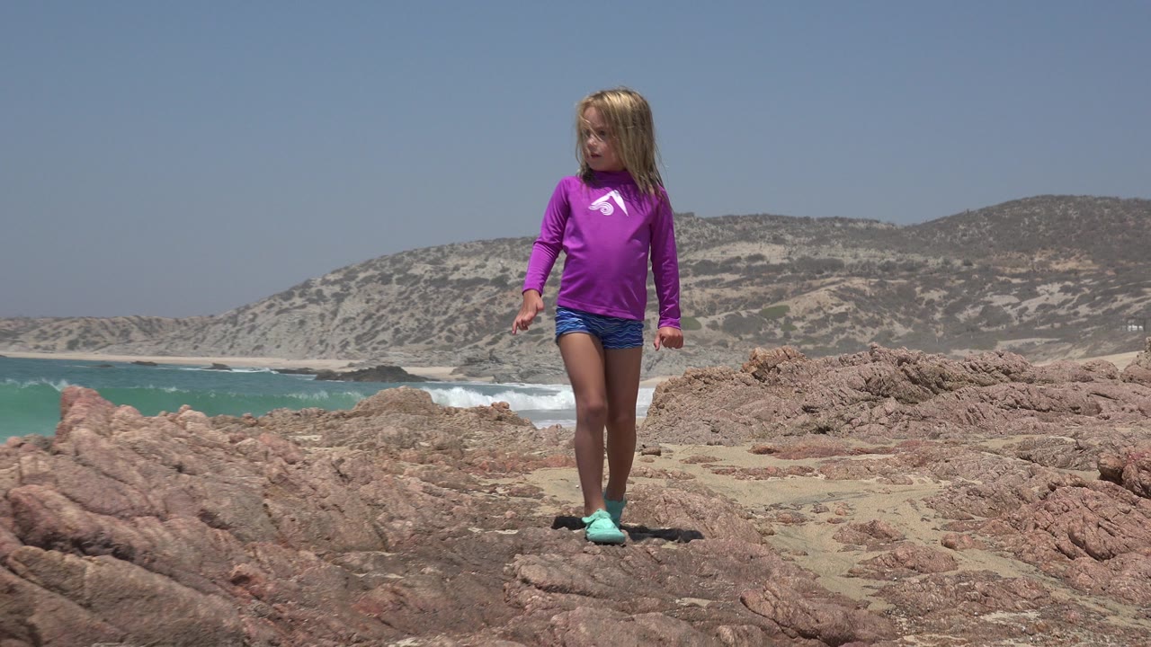 Child in a purple shirt stands farther along the rocky shore in Cabo San Lucas, Mexico, with waves rolling toward a sandy beach below the hills.