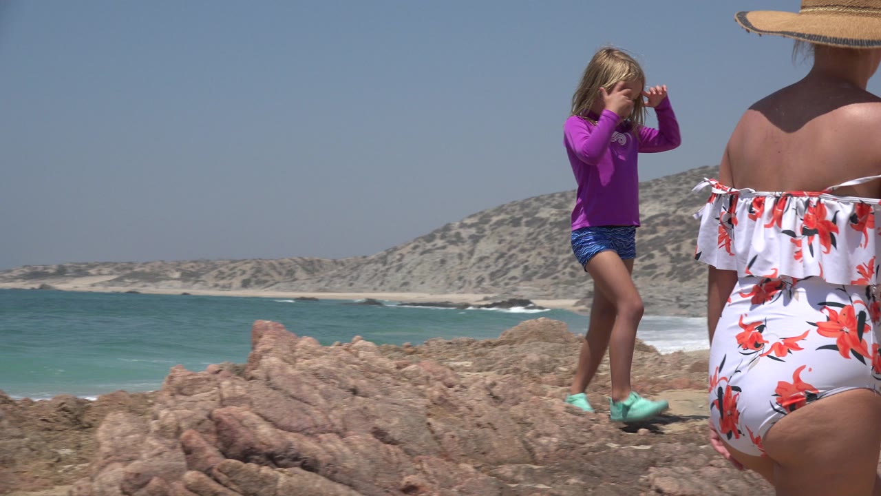 The child remains on the sunlit rocks in Cabo San Lucas while a partially framed adult in a white floral swimsuit stands in the foreground and waves break along the beach behind them.