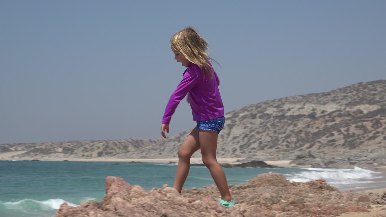 Young girl in purple shirt walking on tan coastal rocks.