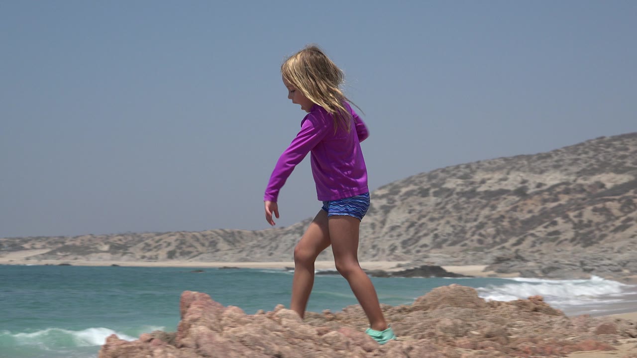 Girl in a purple shirt continues across the tan rock, with blue-green water and low sandy hills filling the Cabo San Lucas background.
