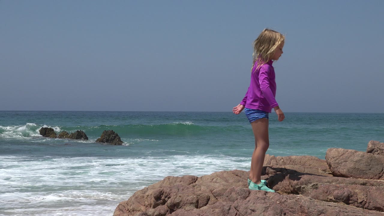 A child in a purple shirt stands on reddish rocks near the sea in Cabo San Lucas, Mexico, with white waves breaking behind them.