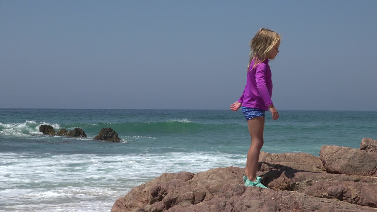 The child begins moving across the rocky foreground in Cabo San Lucas, Mexico, while small waves roll toward the shore behind them.