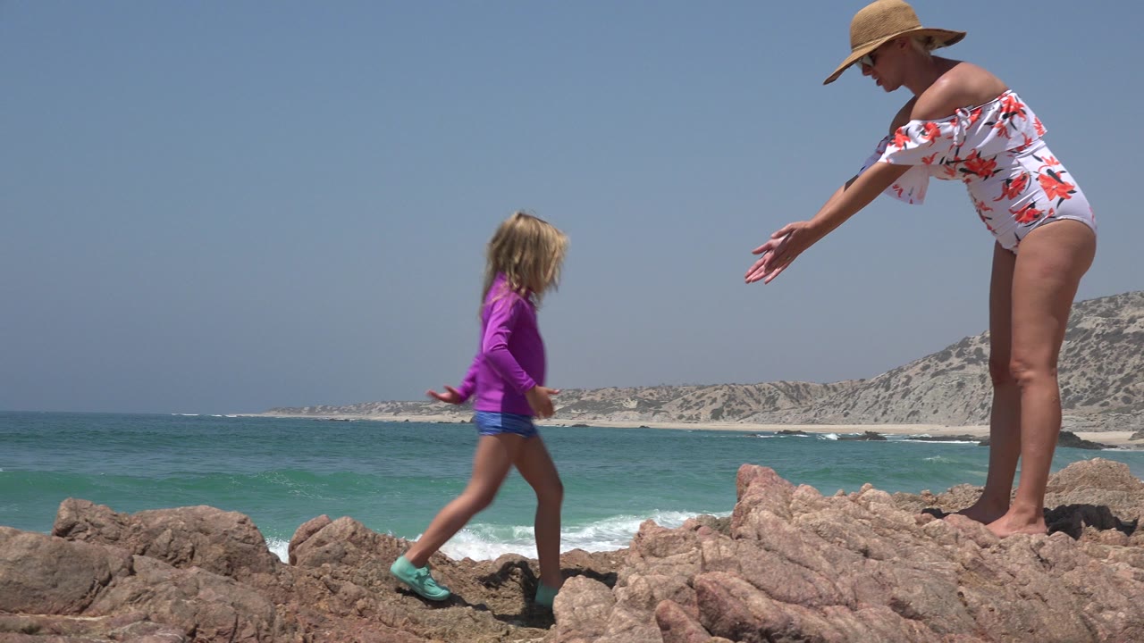Woman reaches toward a young girl on rocks beside the ocean.