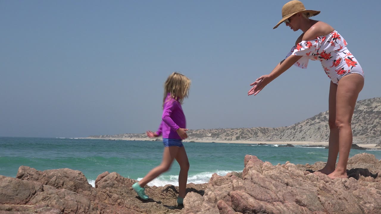 In Cabo San Lucas, Mexico, the young girl steps closer to the woman with her arms partly raised, while surf and pale coastal hills sit behind them.