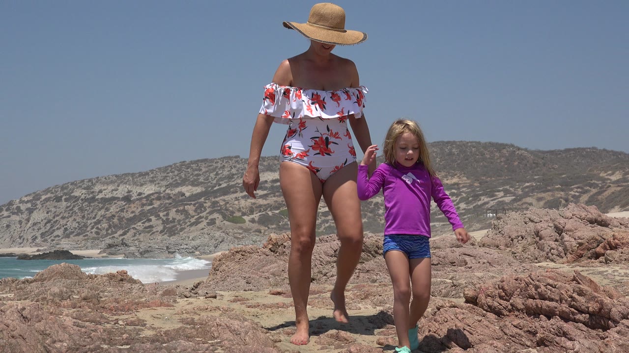 Woman in a straw hat walks with a child across rocks by the beach.