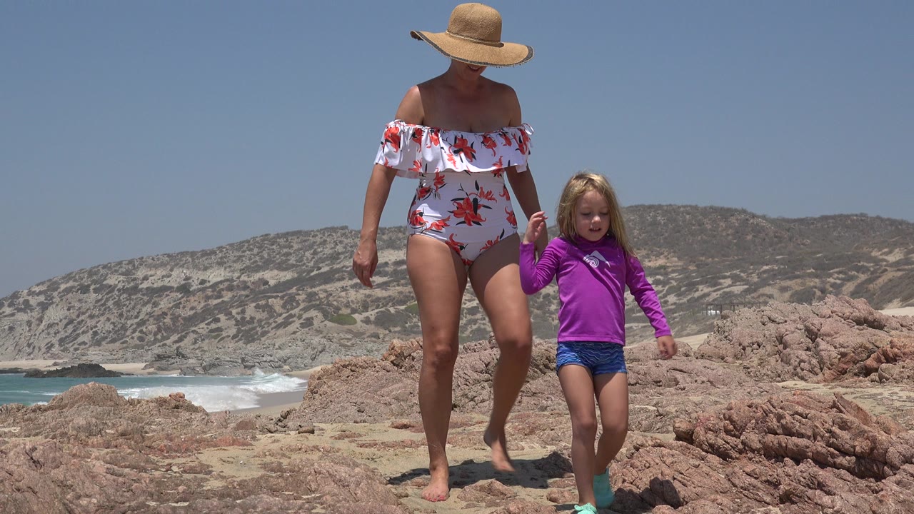 The woman and child remain close together in Cabo San Lucas as they walk over the rocky beach, with a breaking wave visible to the left and dry hills across the horizon.