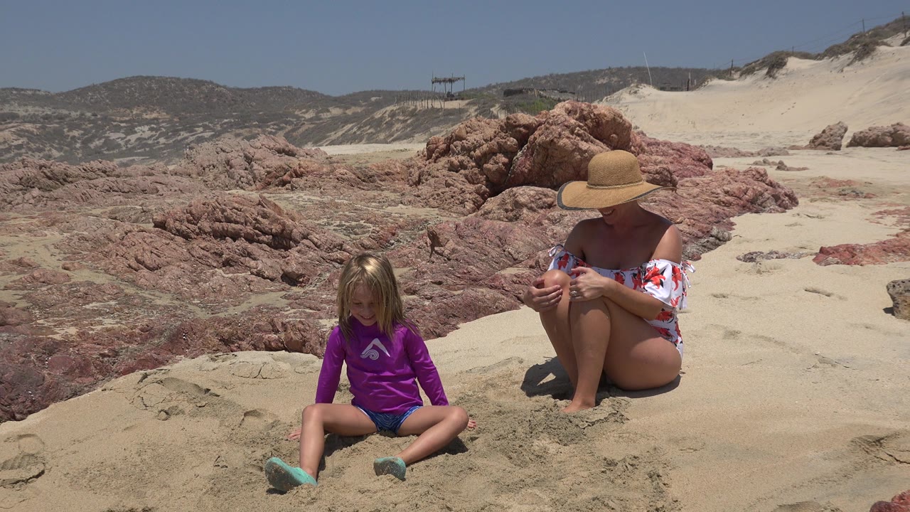 At 0:01 in Cabo San Lucas, Mexico, the child sits facing forward on pale sand as the woman in a wide hat looks downward near the red-toned rocks.