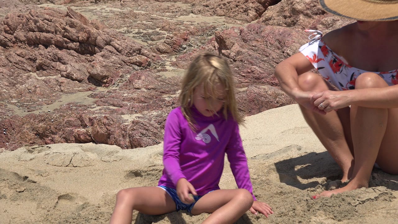 Child in purple shirt sitting on sand near a seated woman