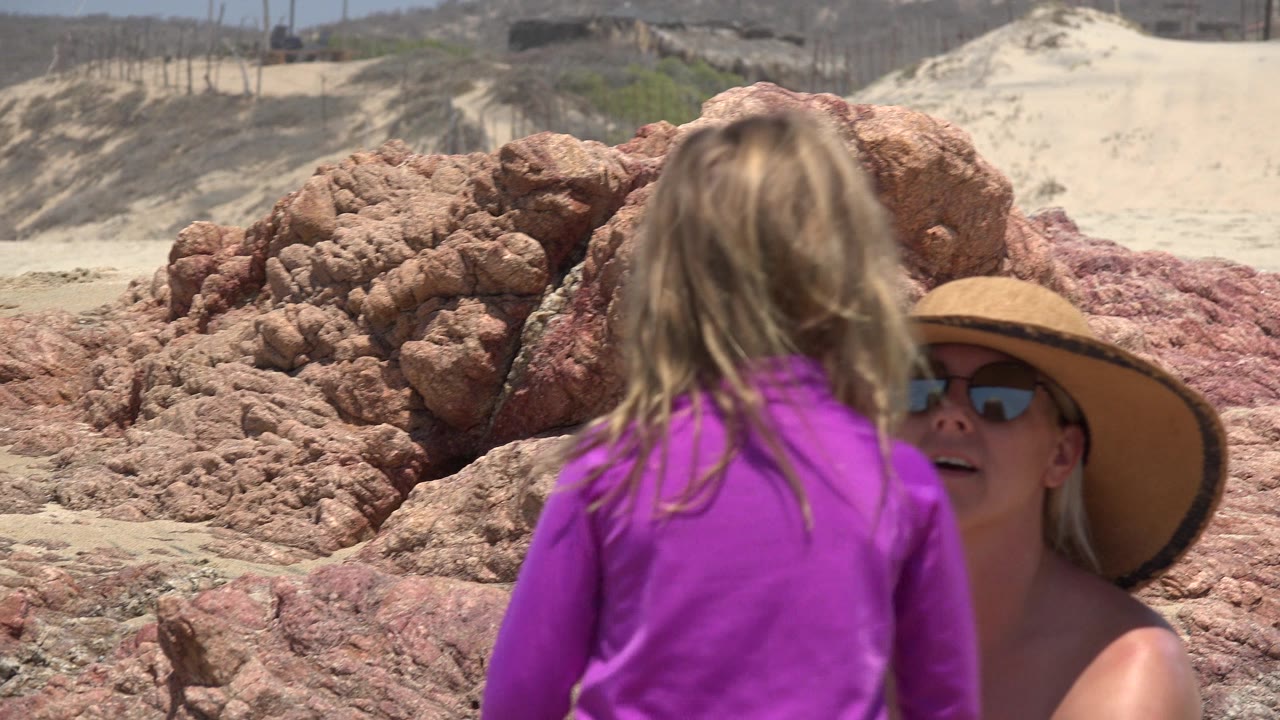 In Cabo San Lucas, a child in a purple rash guard stands with her back to camera while a seated woman in a wide sun hat is partly visible against red coastal rocks.