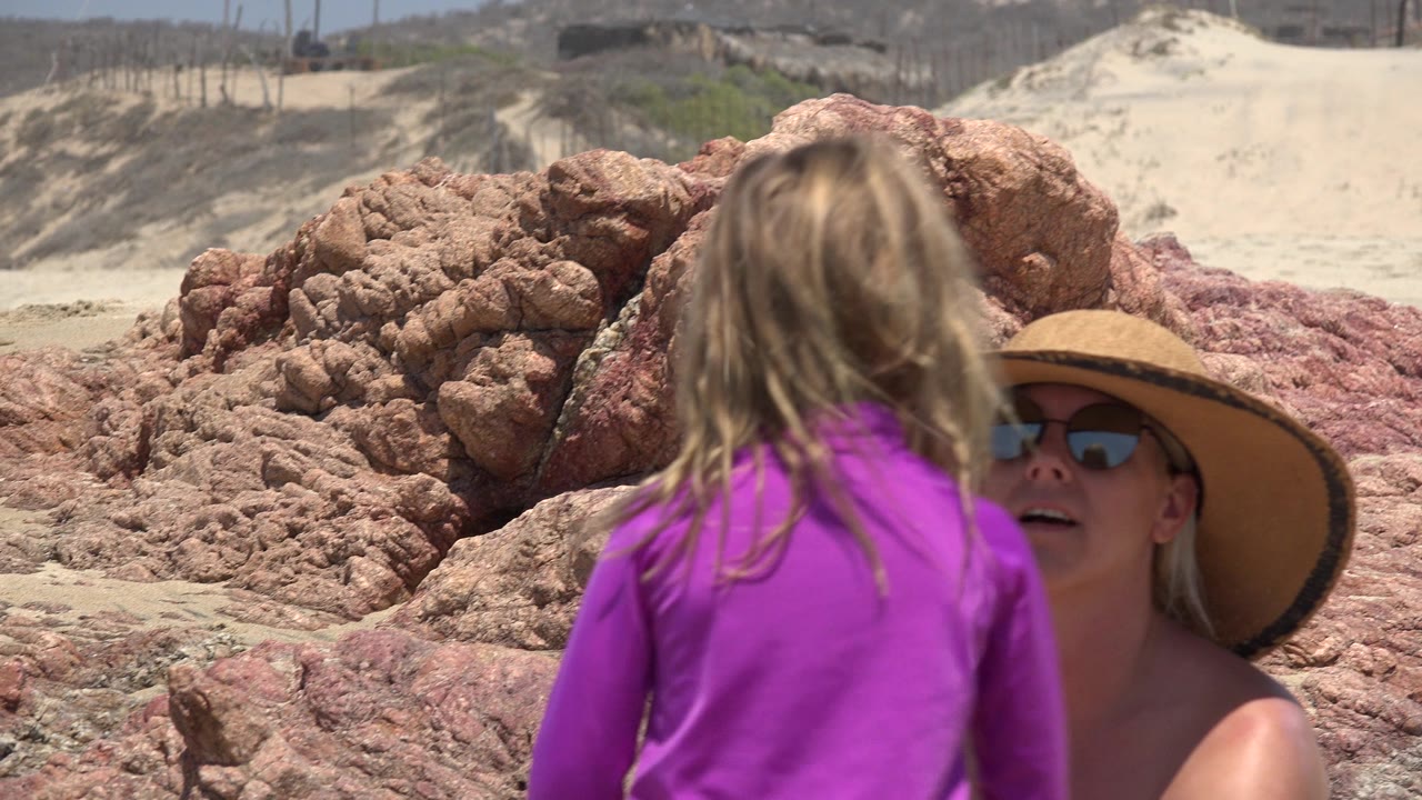 In Cabo San Lucas, the child remains centered in the foreground as the woman in sunglasses and a broad hat looks up from behind her near red rocks and sand.