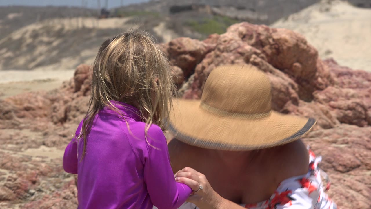 A child in a purple swim shirt sits or crouches beside a woman in a tan sun hat on a rocky beach in Cabo San Lucas, Mexico.