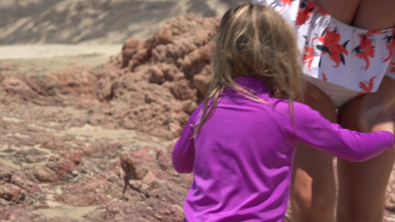The child in a purple swim shirt reaches toward the woman in a white floral swimsuit as they stand near reddish rocks on the beach in Cabo San Lucas, Mexico.