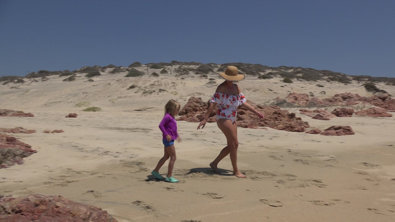 The mother and child continue across the sandy Cabo San Lucas beach, with the child slightly behind and the red rock outcrops stretching across the background.