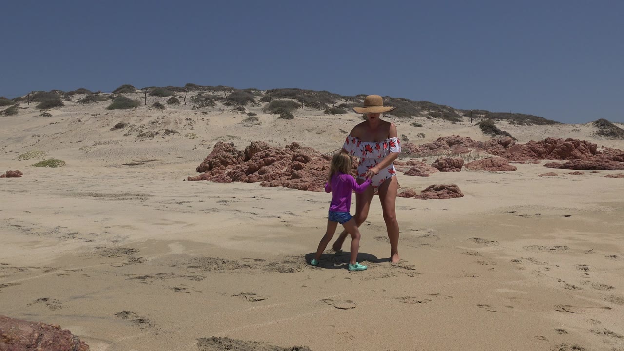 Adult woman and young girl standing on a sandy beach near red rocks.
