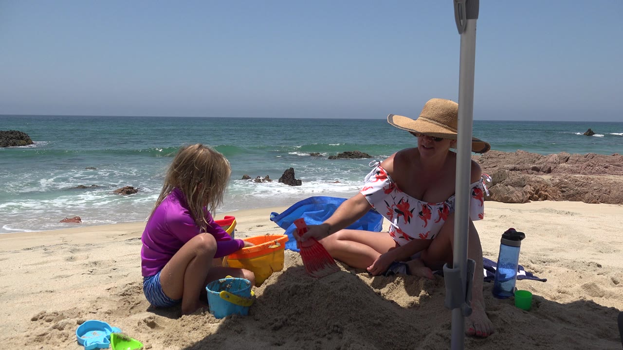 Child reaches toward a bucket beside a woman on the sand