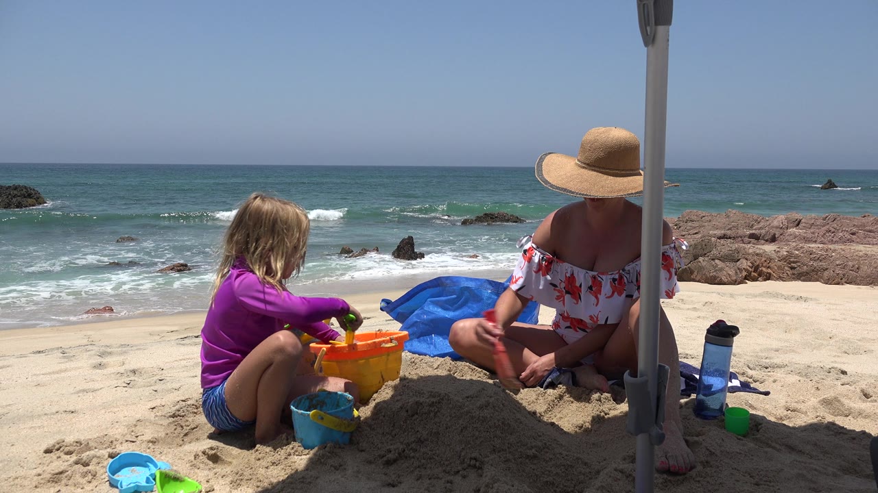 In Cabo San Lucas, Mexico, the child faces the bucket and the woman sits at the edge of the sand pile, with beach toys, a water bottle, surf, and dark rocks visible.