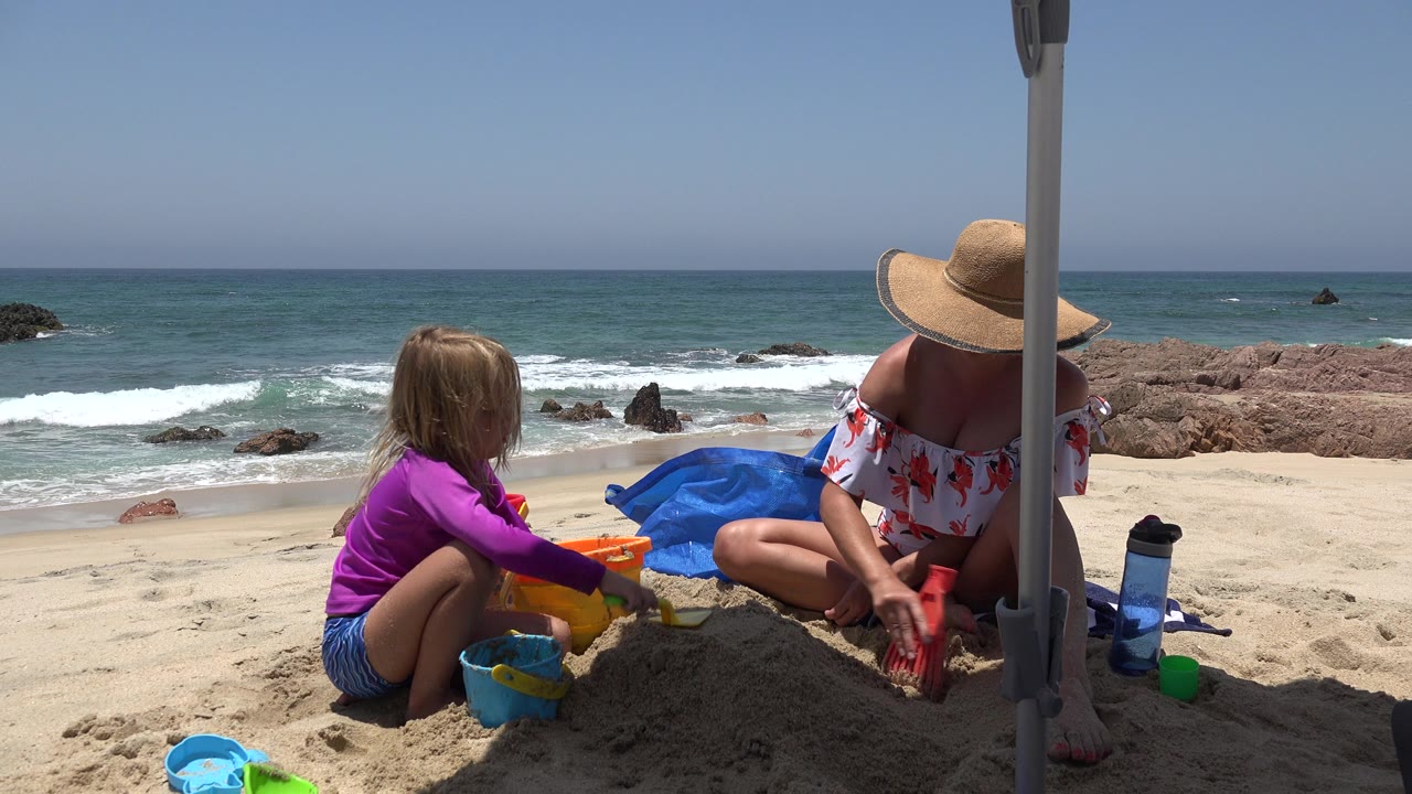 Woman and child sit on a sandy beach with toys as waves break behind them.