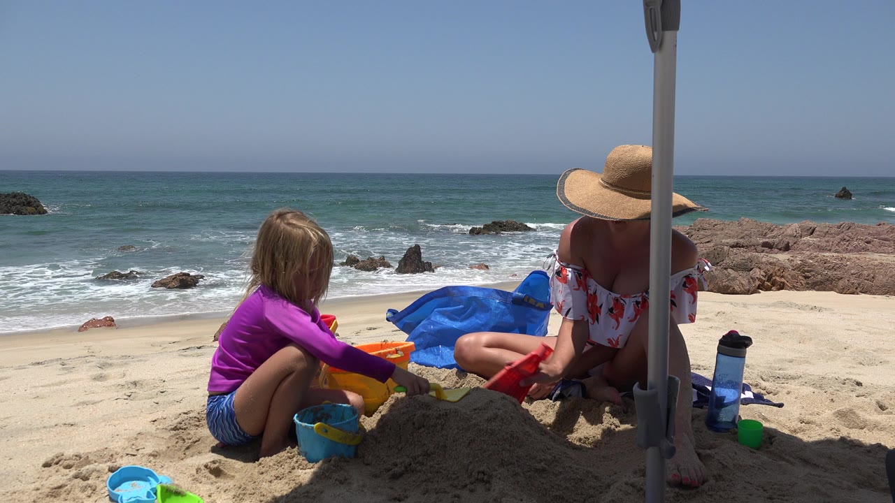 Child and adult sitting on a sandy beach with toys near the ocean.