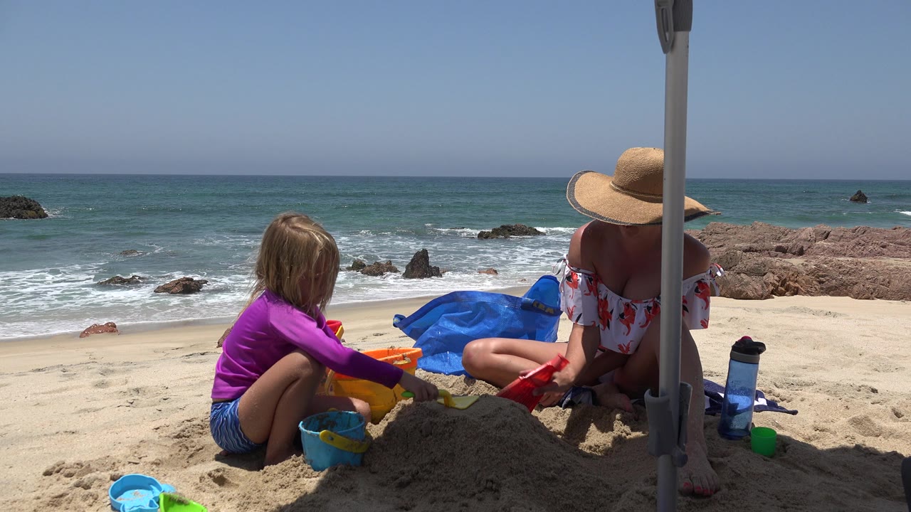 The girl leans toward the sand toys as the adult sits close by under the umbrella pole on the Cabo San Lucas beach, with small waves rolling behind them.