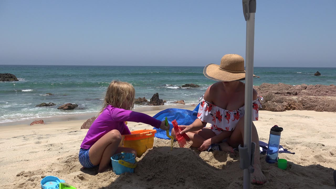 At 0:00 in Cabo San Lucas, a child in a purple top and an adult in a straw hat sit beside buckets and shovels while ocean waves and rocks fill the background.