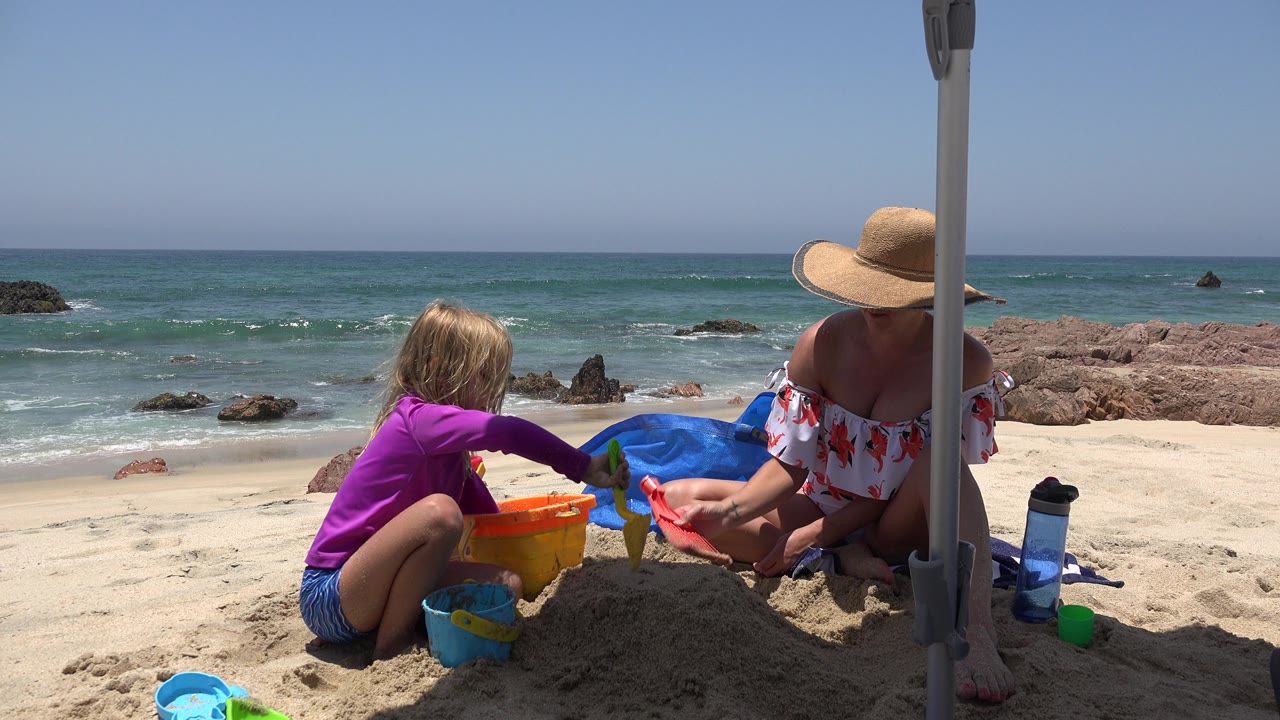 Child and adult continue building in the sand with ocean and rocks behind them.