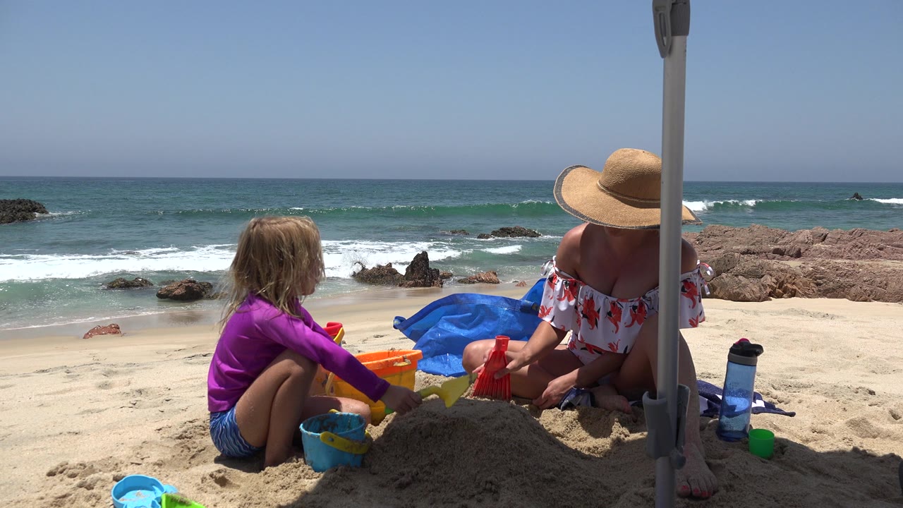 At 0:00 in Cabo San Lucas, a child in a purple top and an adult in a straw hat sit under an umbrella with buckets and shovels as waves break behind them.