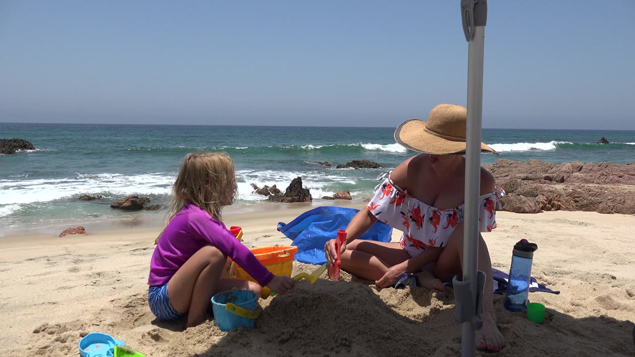 Adult holds a red shovel while child plays with buckets on the sand.
