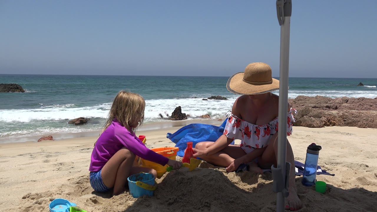 A child in a purple top and an adult woman in a straw hat sit on a Cabo San Lucas beach with colorful sand toys, waves, rocks, and blue ocean behind them.
