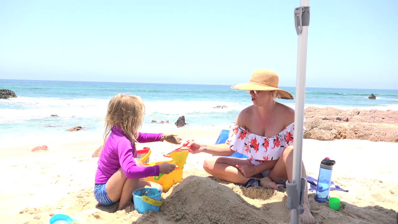 At 0:01 in Cabo San Lucas, Mexico, the child extends an arm toward the seated woman while a yellow bucket, blue bucket, and red toy sit on the bright sand.
