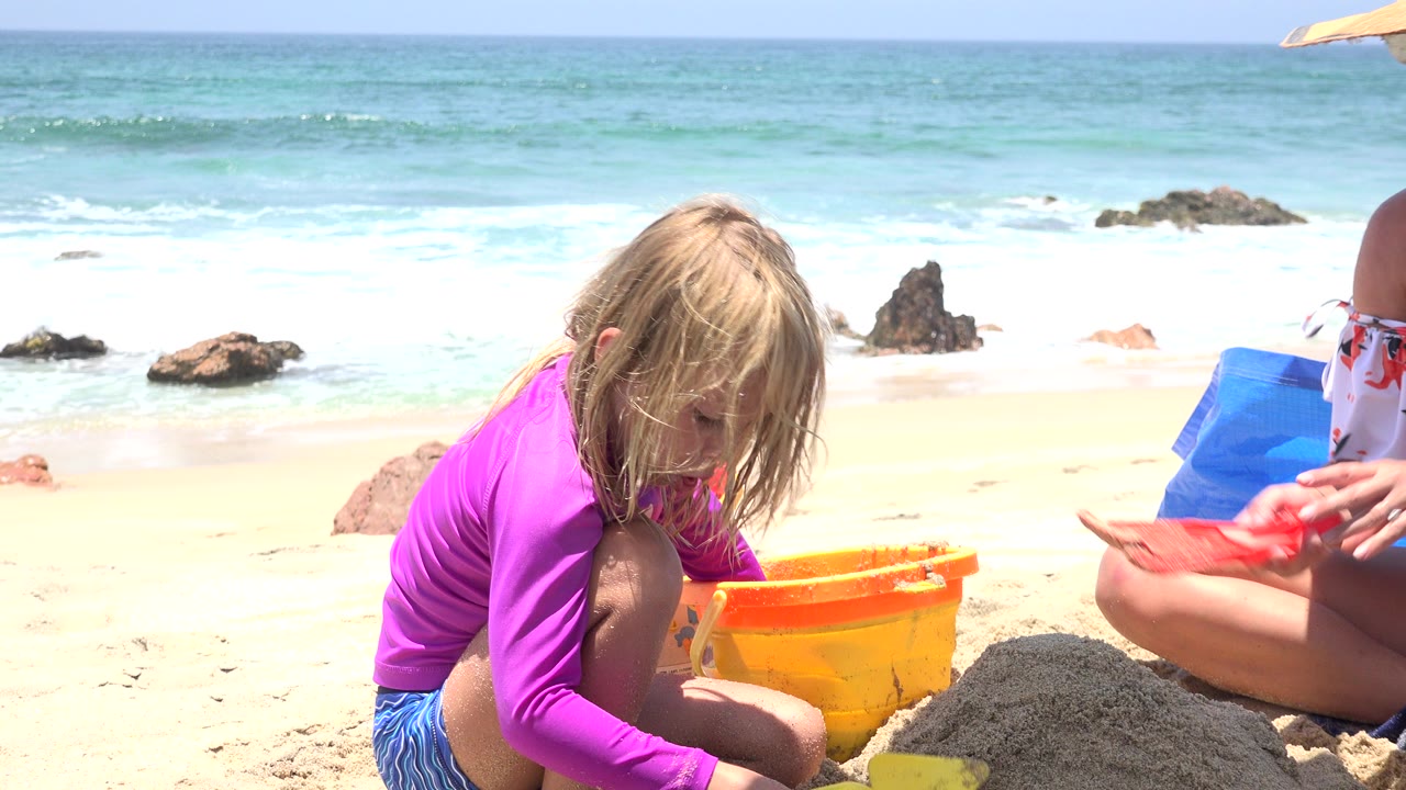 Young girl in pink playing in sand beside an orange bucket at the beach.