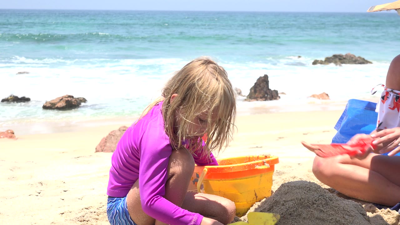 At 0:01, the girl stays low beside the orange bucket as a red sand shovel held by a partly visible seated person appears at the right edge of the Cabo San Lucas beach frame.