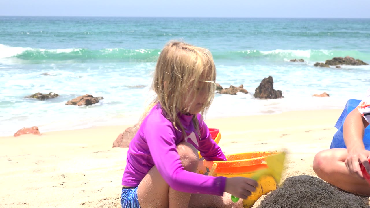 Blonde child in a purple swim shirt plays with sand toys on a beach.