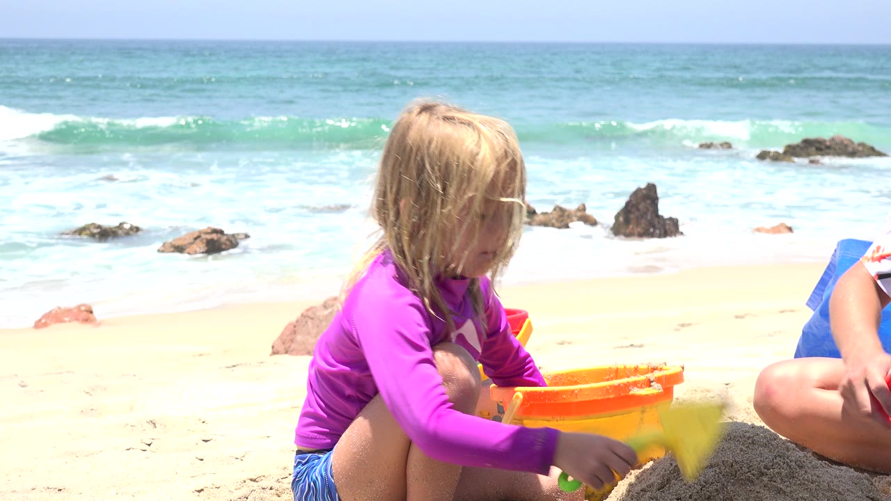 At 0:01 in Cabo San Lucas, the child remains crouched near the orange bucket and moves the yellow shovel over a sand mound, with foamy waves rolling behind her.