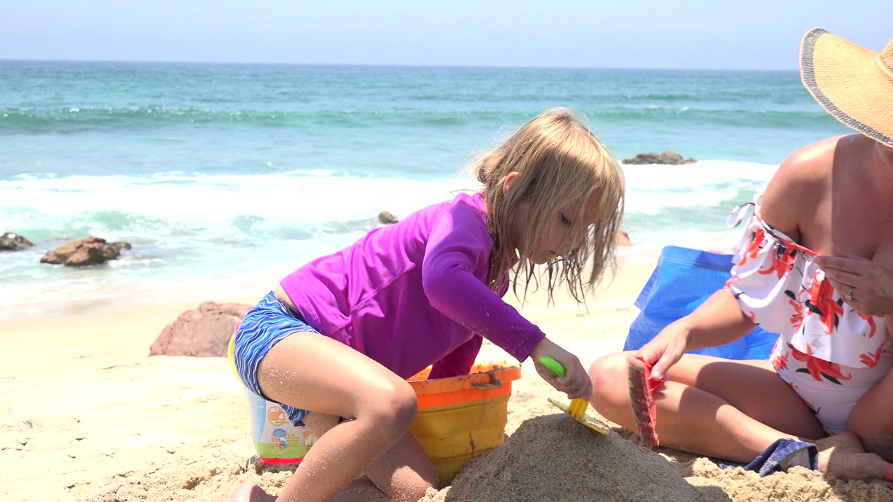 The child bends lower over the sand mound with a green-handled toy while the woman holds a red mold near the orange bucket on the Cabo San Lucas shoreline.