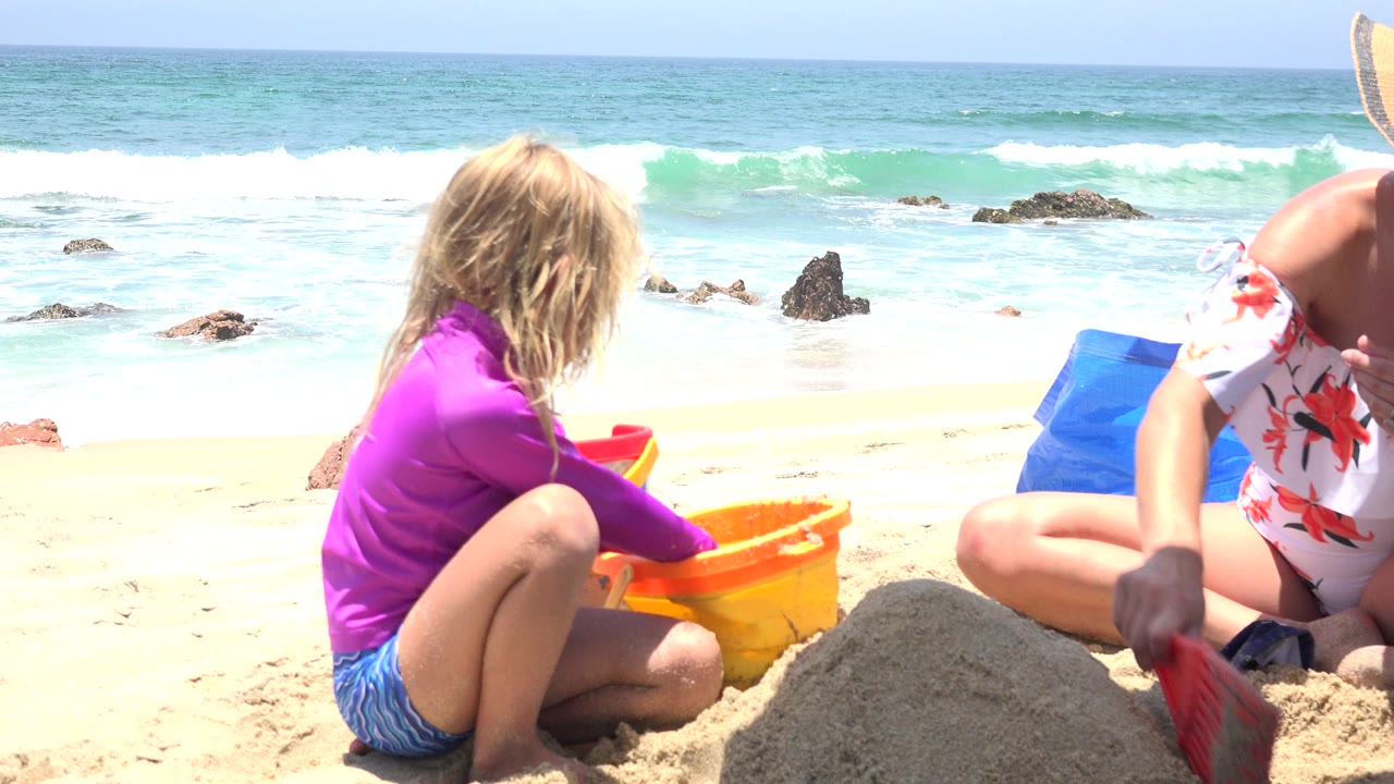 A child in a purple shirt sits in the sand beside a yellow beach bucket in Cabo San Lucas, Mexico, with surf, rocks, and a partly visible person at the right edge.
