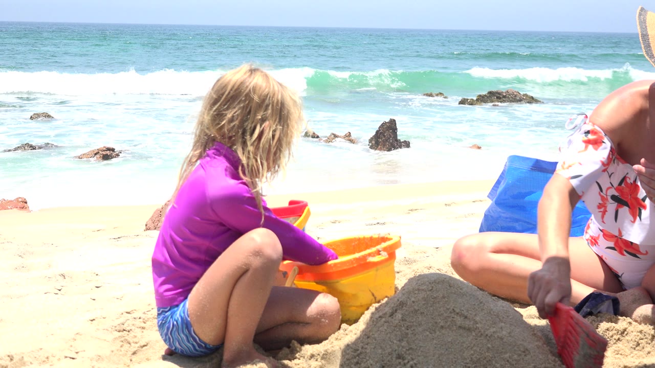 The child in the purple shirt leans near the yellow bucket as a partly visible person reaches in with a red shovel, with waves and dark rocks behind them in Cabo San Lucas, Mexico.
