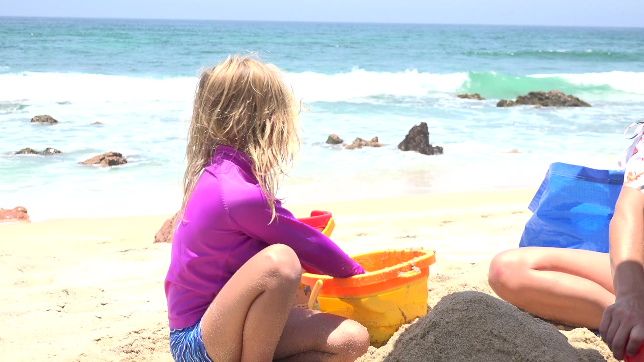 Child sits by a yellow bucket as surf breaks behind the beach.
