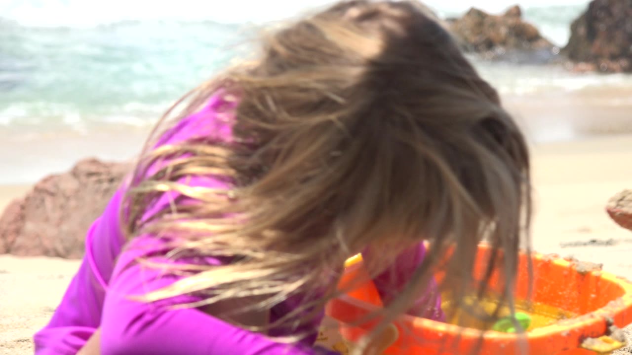 A child in a purple top bends low over an orange beach bucket on white sand in Cabo San Lucas, with pale surf and dark rocks behind her.